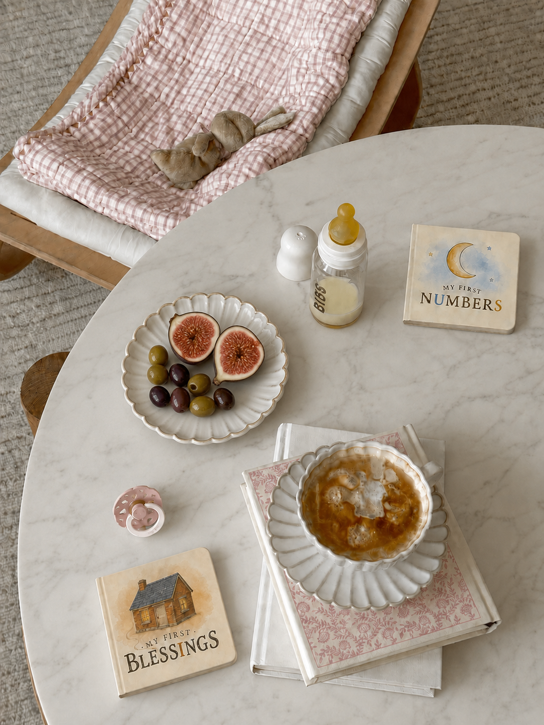 A styled tabletop scene with a white marble round table holding a cup of iced coffee on a scalloped saucer, a plate of sliced figs and olives, a baby bottle, two decorative books or cards reading 'My First Numbers' and 'My First Blessings,' and a pink gingham blanket draped over a wooden chair in the background.